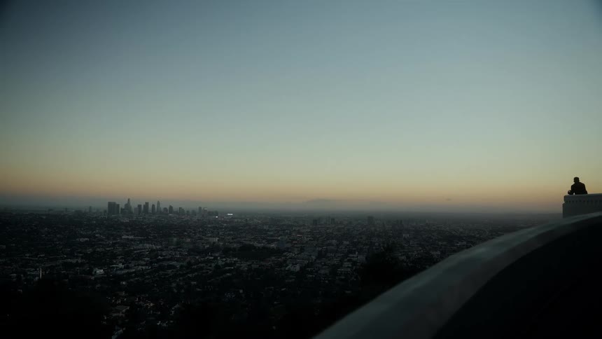 A lone silhouette watches the vast Los Angeles cityscape from a high vantage point at dusk