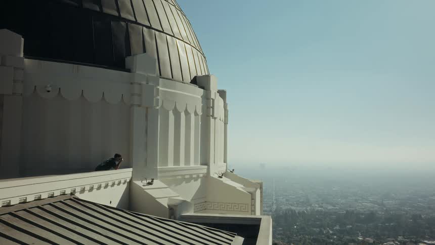 A man walks along the balcony of the Griffith Observatory before stopping to admire the vast, hazy cityscape of Los Angeles