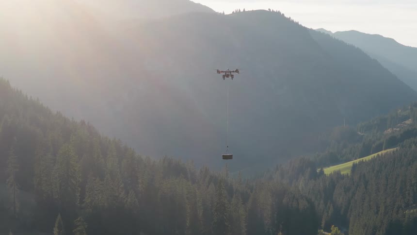 A heavy-lift cargo drone transports a package on a long line over a vast, forested mountain valley during a hazy sunrise