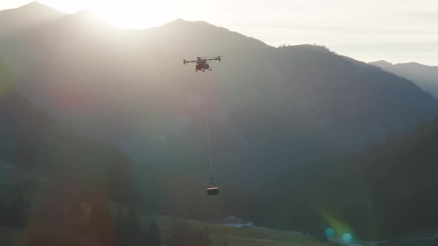 A heavy-lift drone transports a payload on a cable over a forested mountain valley at sunrise