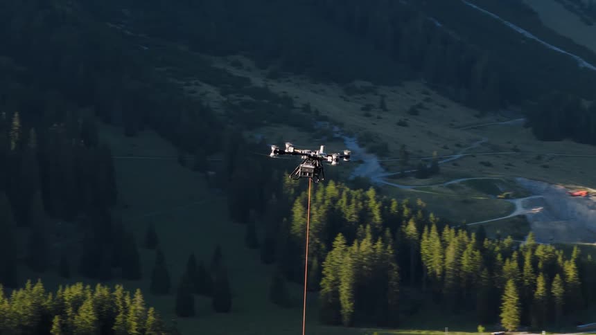 A large octocopter drone carrying a long red hose flies over a forested mountain valley