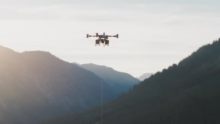 A heavy-lift industrial drone carrying cargo on a long rope flies out of frame to reveal a beautiful mountain valley at sunrise
