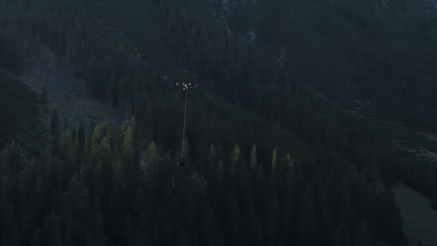 A heavy-lift cargo drone transports a package on a longline over a dense, mountainous forest at dusk