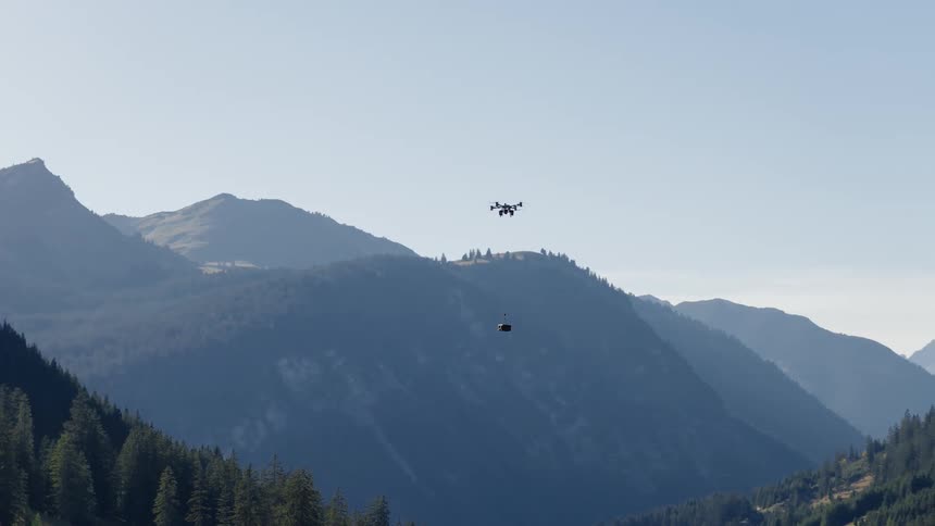 A large industrial drone carries a payload container while flying over a vast, forested mountain valley