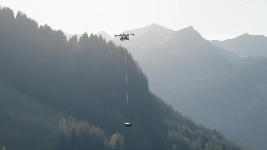 A heavy-lift cargo drone with a suspended payload ascends over a lush forest, revealing a majestic mountain range in the background