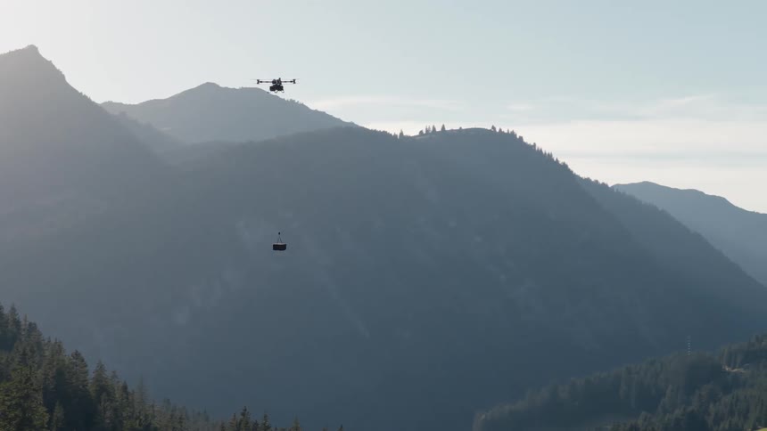 A heavy-lift cargo drone flies across a vast mountain valley, lowering its suspended package for delivery