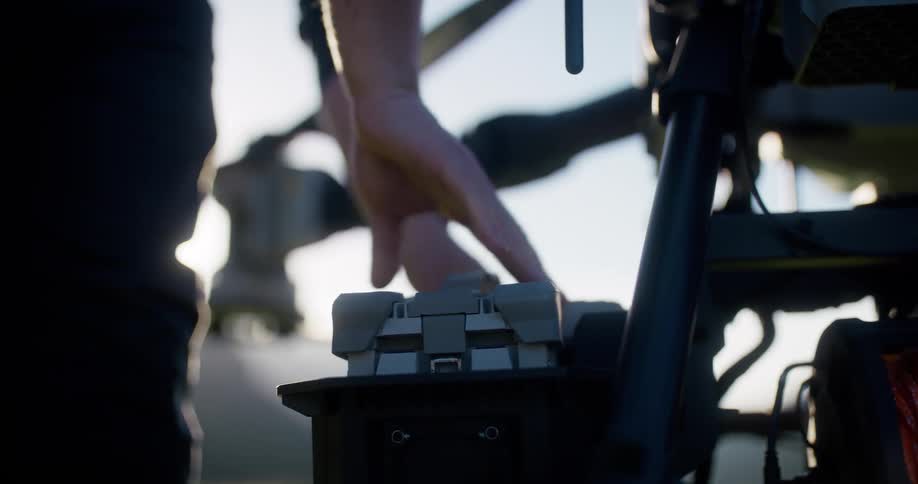 A close-up slow-motion shot shows a person inserting a large battery into a professional drone as the sun flares in the background