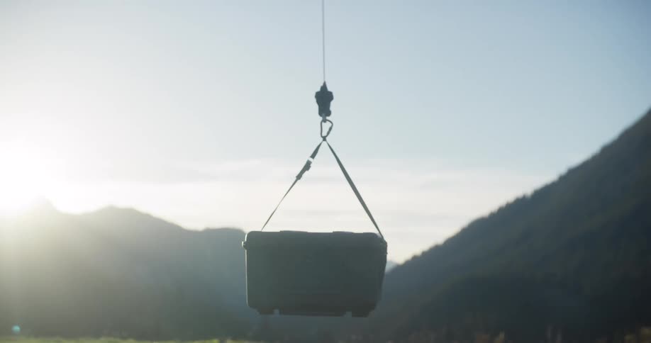 A hard-sided equipment case is lifted by a hoist from a green field against a stunning mountain backdrop at sunrise
