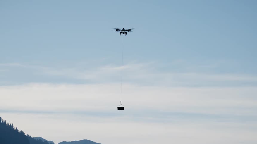 A silhouette of a heavy-lift cargo drone transports a package on a long line while flying through the sky