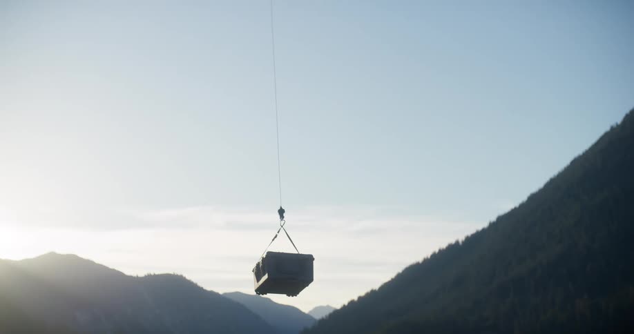 A heavy-duty industrial drone lifts a rugged cargo case from a grassy field and flies away over a beautiful mountain valley
