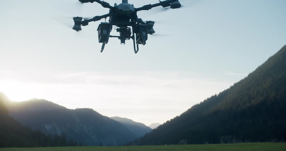 Slow motion shot of a large professional camera drone descending to hover over a green meadow in a mountain valley at sunset