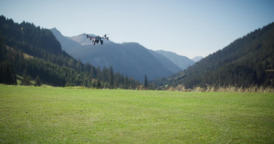 A professional-grade octocopter drone takes off from a lush green meadow and flies away towards a stunning mountain valley on a sunny day