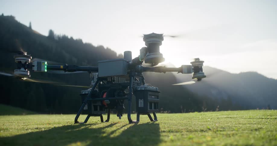 A professional drone takes off in slow motion from a grassy field in the mountains, silhouetted against the bright sun