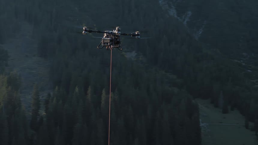 A heavy-lift drone transports a payload on a long line over a dense forest in a mountain valley