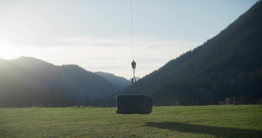 A rugged black case is lowered by a cable and gently lands on a grassy field with a beautiful mountain landscape in the background
