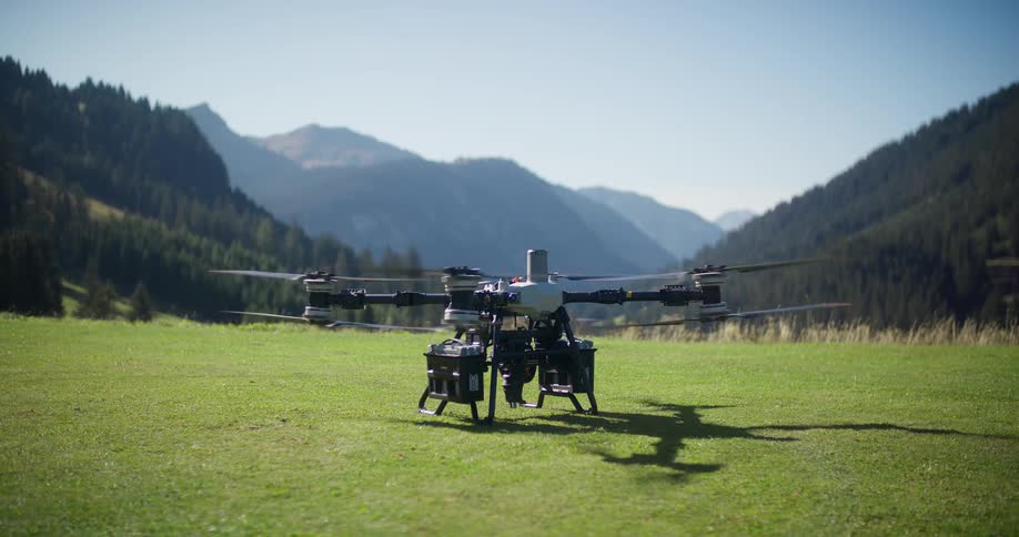 A large, industrial heavy-lift drone takes off in slow motion from a grassy meadow in a beautiful mountain valley on a sunny day