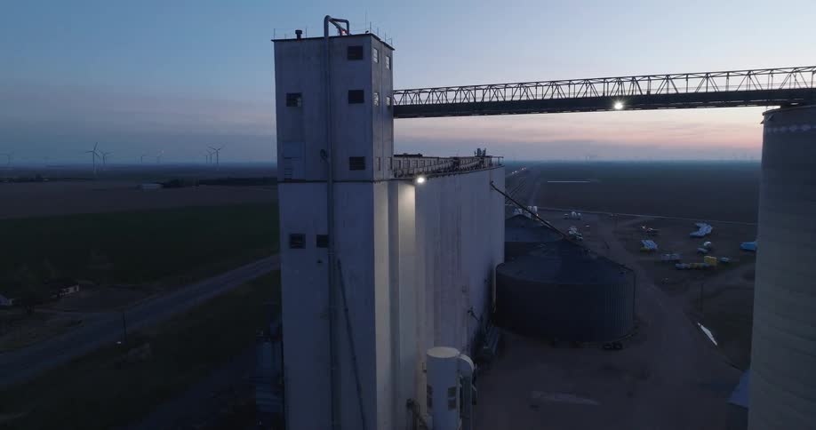 A serene aerial flyover of a towering grain elevator complex in the American heartland at twilight, with wind turbines dotting the horizon