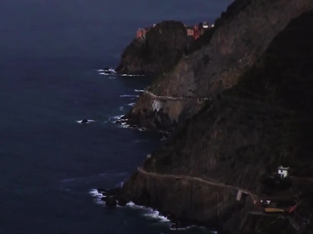 Dusk descends on the dramatic Italian coastline, showing the famous coastal path winding along the cliffs towards a village