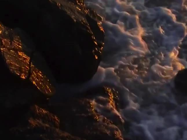 Golden light from a sunrise glistens on waves as they slowly wash over dark coastal rocks