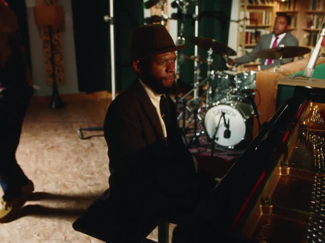 A stylish Black man in a hat plays a grand piano with passion while his band performs in a cozy music studio