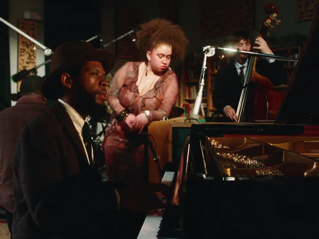 A stylish jazz trio with a pianist, a female vocalist, and a bassist performs a soulful song in a dimly lit studio