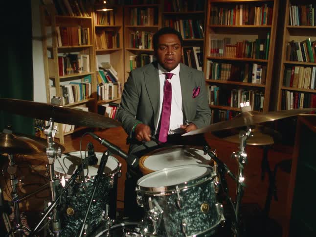 A passionate African American musician in a suit plays a dynamic jazz solo on the drums in a cozy, book-lined studio