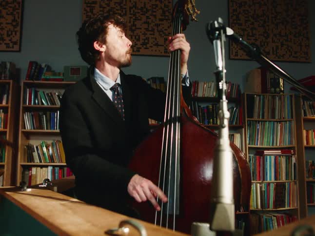 A serious musician in a suit plays the double bass during a recording session in a cozy studio with bookshelves