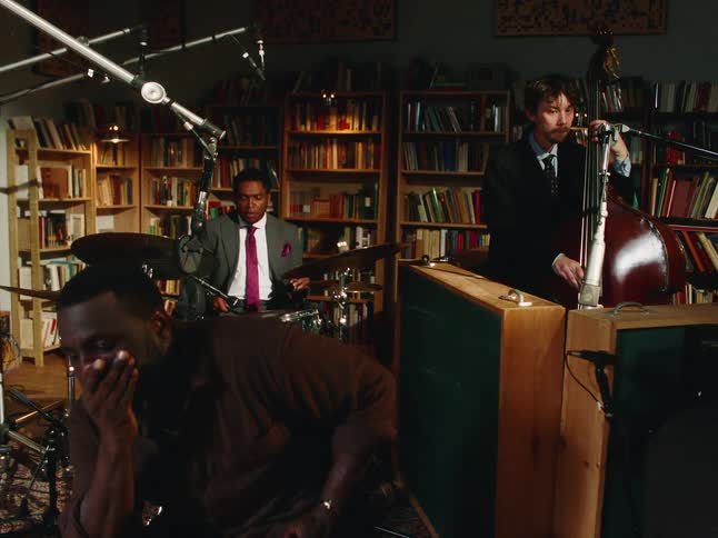 A jazz trio performs a soulful song in a cozy, book-lined studio while another man listens intently