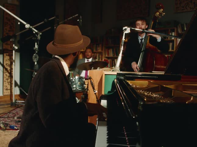 A stylish jazz trio with a pianist, bassist, and drummer performs a soulful song in a cozy, intimate studio