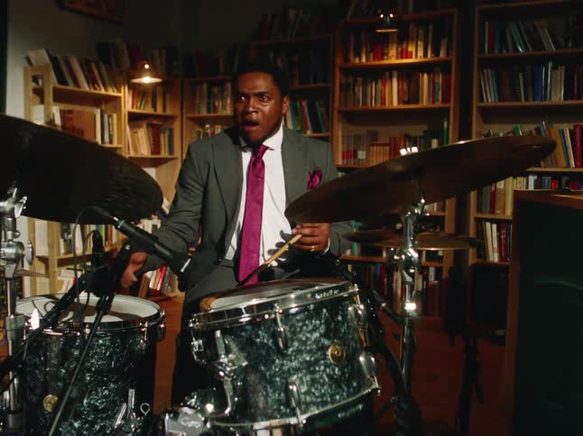 An expressive jazz drummer in a suit plays an energetic solo in a cozy, book-lined studio as the camera slowly pushes in