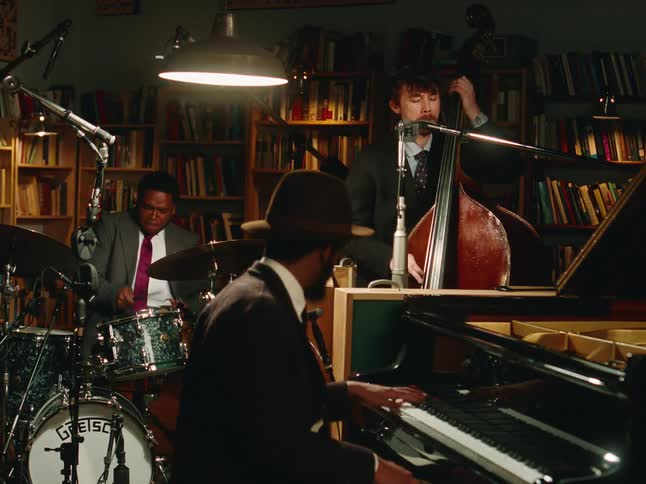 A stylish jazz trio passionately performs on piano, upright bass, and drums in a cozy, dimly lit studio surrounded by books