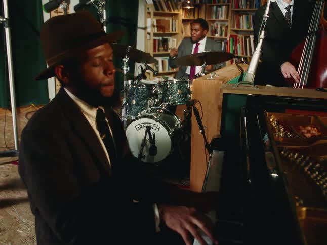 A charismatic pianist in a brown hat passionately plays a grand piano while his jazz band performs with energy in an intimate studio filled with books