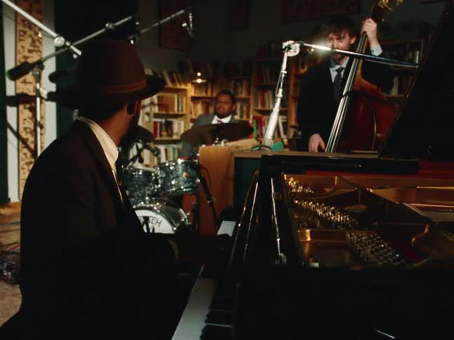 A stylish jazz trio with a pianist, drummer, and bassist performs together in a cozy recording studio