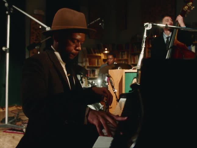 A jazz trio performs in a warm, intimate setting, with a close-up on the pianist in a fedora playing with soul