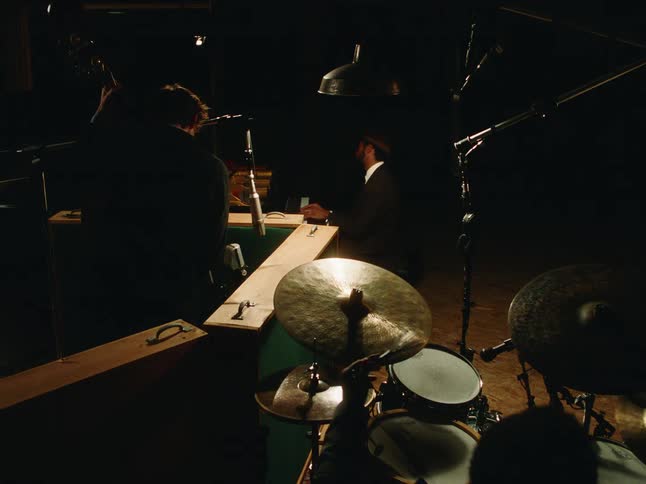 A jazz trio with a pianist, upright bassist, and drummer performs together in a moody, dimly lit recording studio