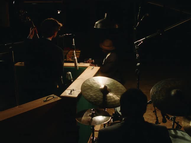 A jazz trio with a pianist, bassist, and drummer performs together in a dimly lit, moody recording studio