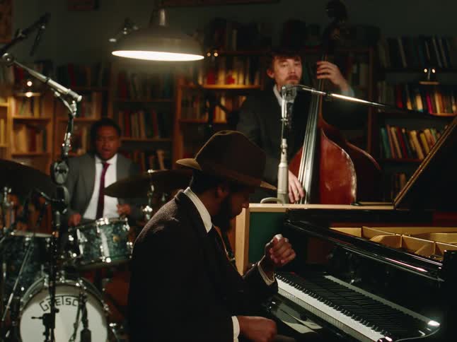 A jazz trio with a pianist, bassist, and drummer performs a soulful song in a cozy, intimate library setting