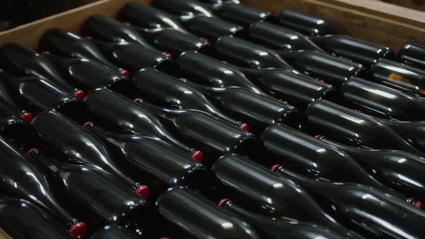 A close-up shot shows a wooden crate filled with rows of dark glass wine bottles with red caps