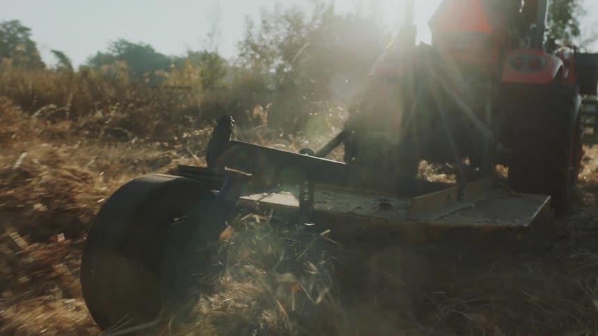 A tractor's mower attachment churns through a dry field in slow motion, backlit by the warm afternoon sun