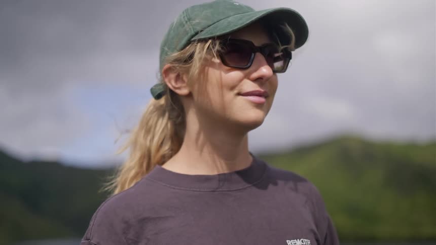 A slow-motion close-up of a young woman in a cap and sunglasses smiling serenely outdoors with mountains in the background