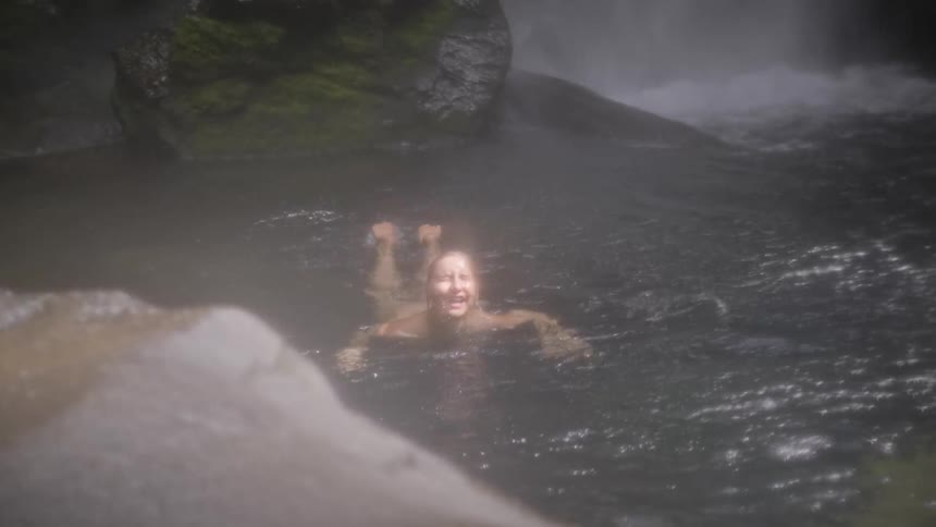 Slow motion of a smiling woman swimming on her back in a misty pool at the base of a waterfall