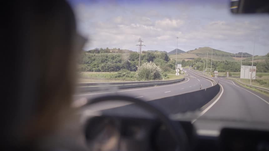 An over-the-shoulder perspective from a car driving along a scenic highway through rolling green hills on a bright day