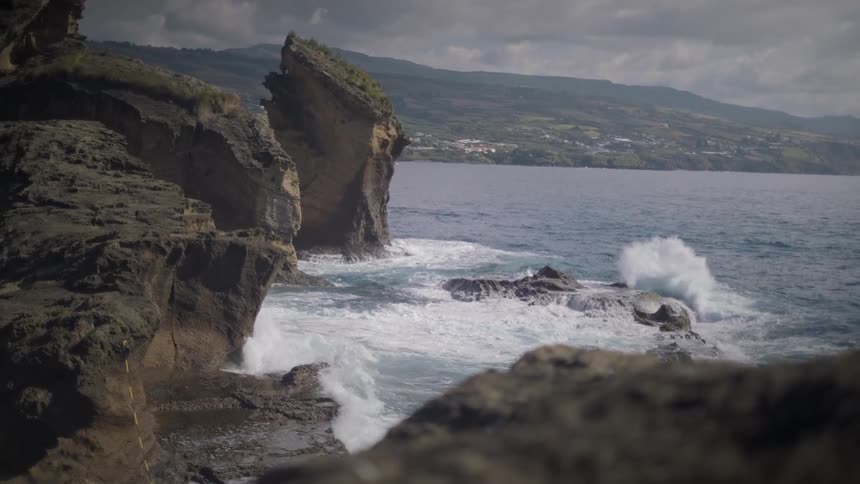 Dramatic slow motion view of powerful ocean waves crashing against a rugged, rocky coastline under an overcast sky