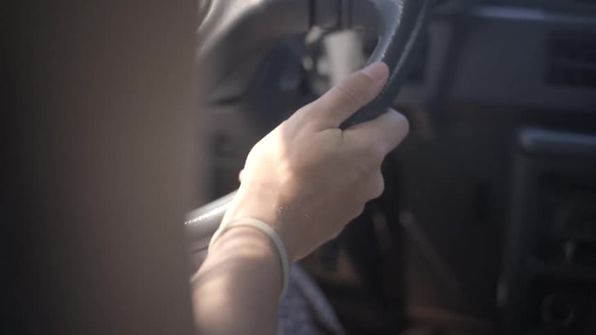 A close-up over-the-shoulder shot shows a person's hand holding the steering wheel while driving a car