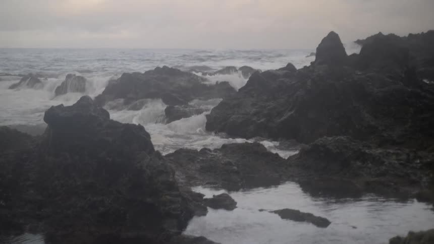 A slow-motion pan reveals ocean waves crashing over a dark, rocky shore and creating small waterfalls into tide pools on a foggy day