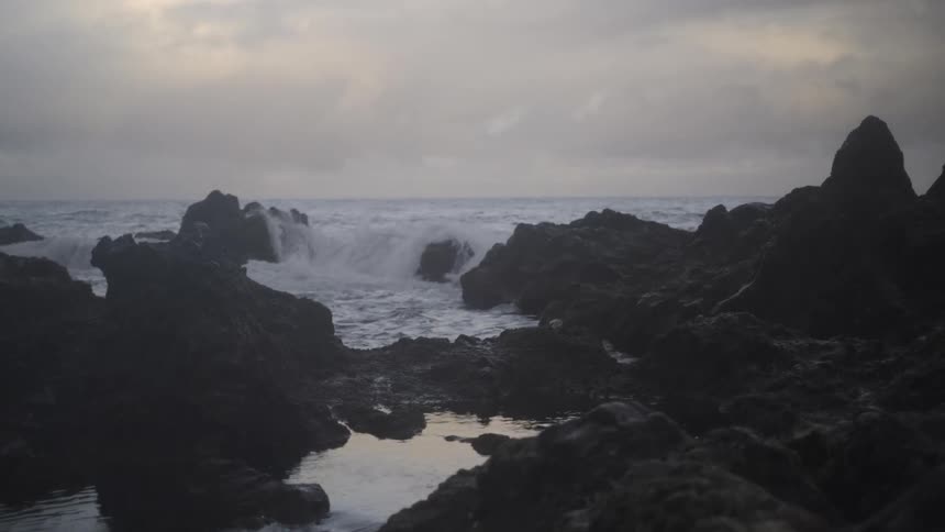 Slow motion shot of powerful ocean waves crashing against a dark, rocky coastline under an overcast sky