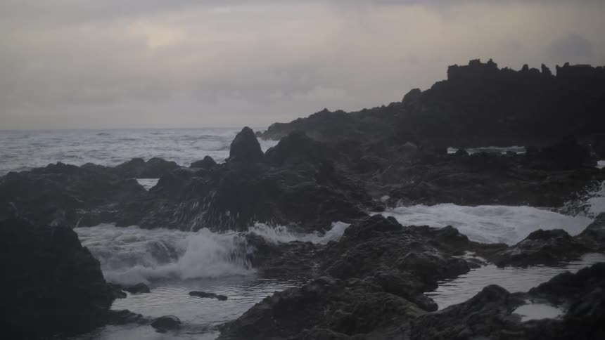 Ocean waves crash in slow motion over jagged volcanic rocks on a gloomy, overcast day