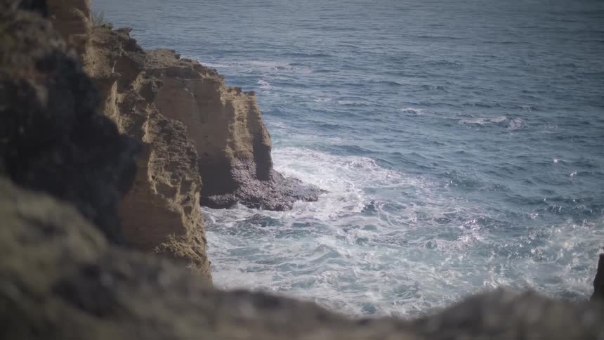 Powerful ocean waves crash against the base of a rugged cliff as seen from a high vantage point