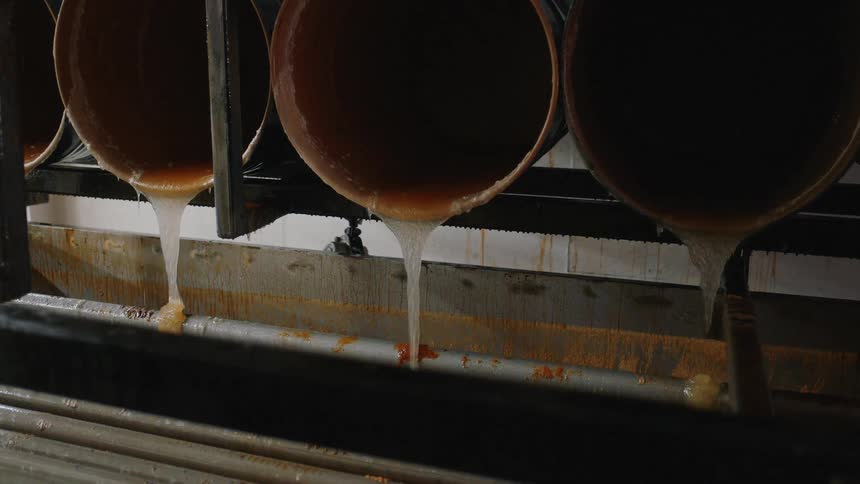 Thick, viscous liquid pours from large industrial vats in a slow, steady stream on a factory production line