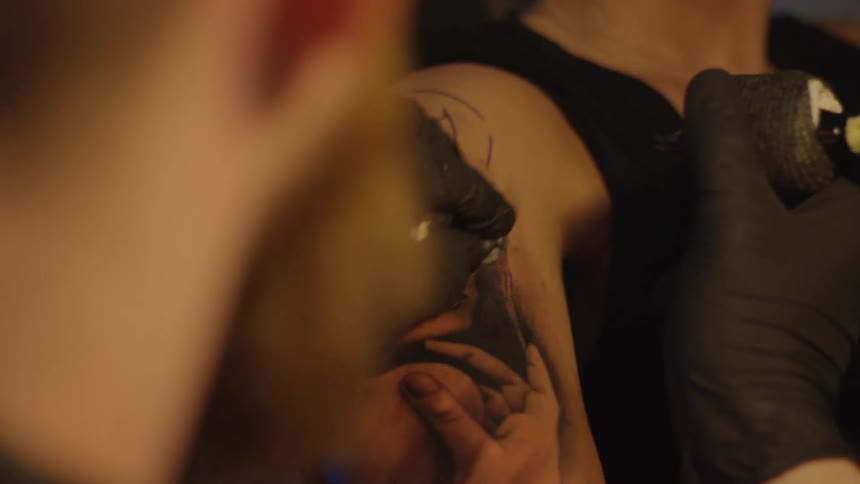 Skilled tattoo artist wearing black gloves meticulously inks a portrait onto a woman's upper arm